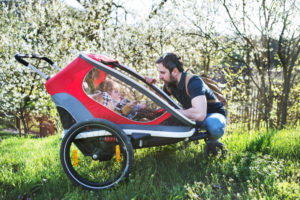 A father looking at two toddler children sitting in jogging stroller outside in spring nature.